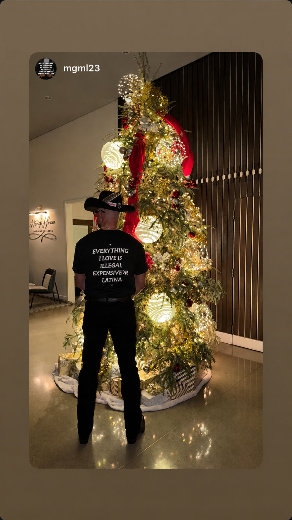 Mand i sort EVERYTHING I LOVE IS ILLEGAL, EXPENSIVE OR LATINA T-shirt ved et oplyst juletræ.