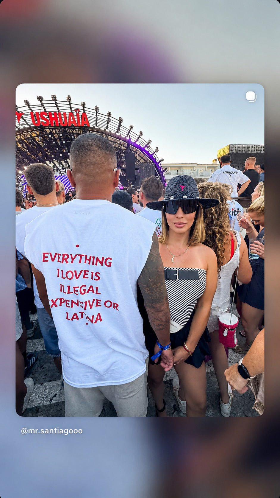 Mand i hvid EVERYTHING I LOVE IS ILLEGAL, EXPENSIVE OR LATINA T-shirt ved en festivalscene.
