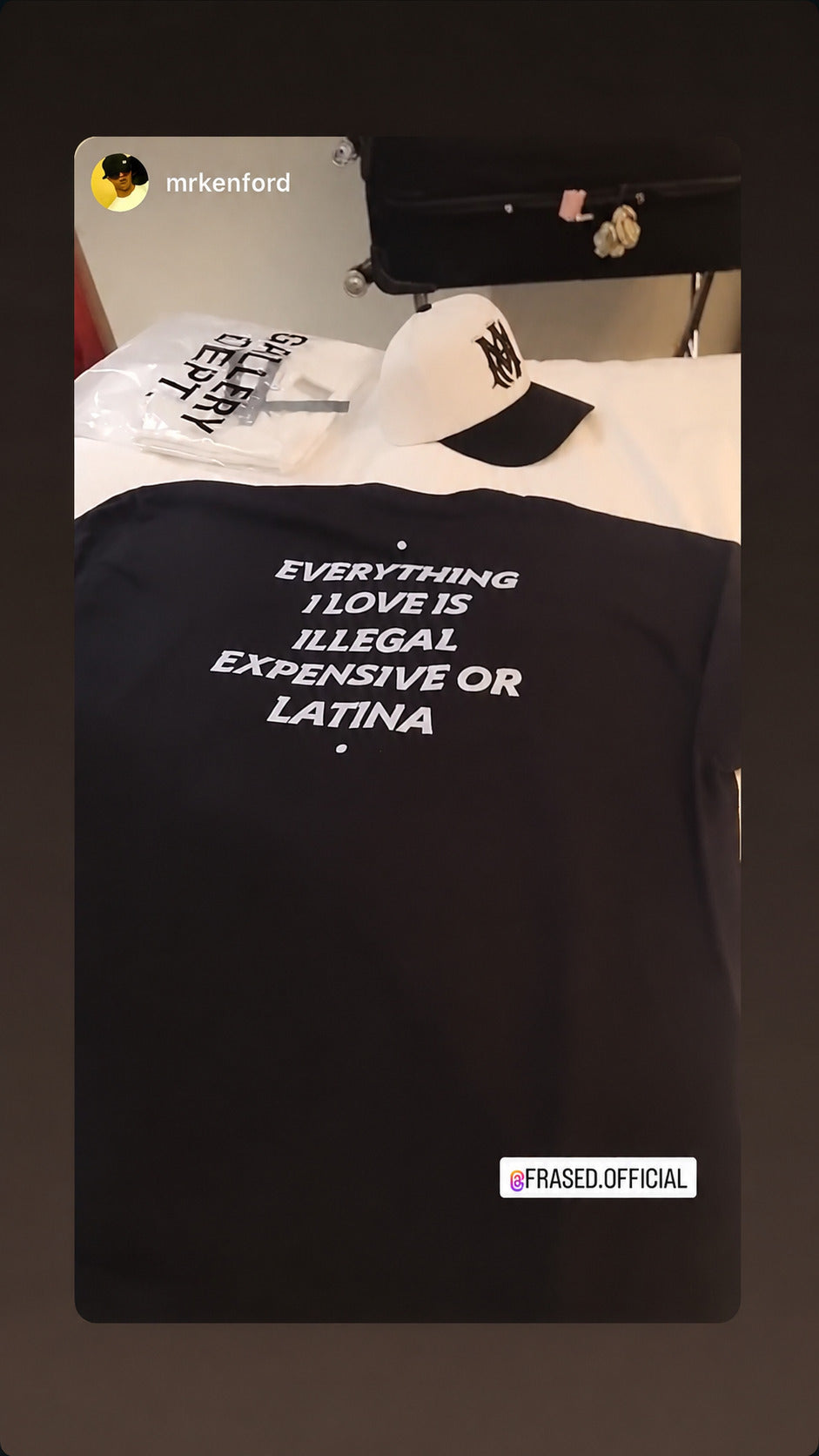 Sort EVERYTHING I LOVE IS ILLEGAL, EXPENSIVE OR LATINA T-shirt lagt frem med kasket på en seng.