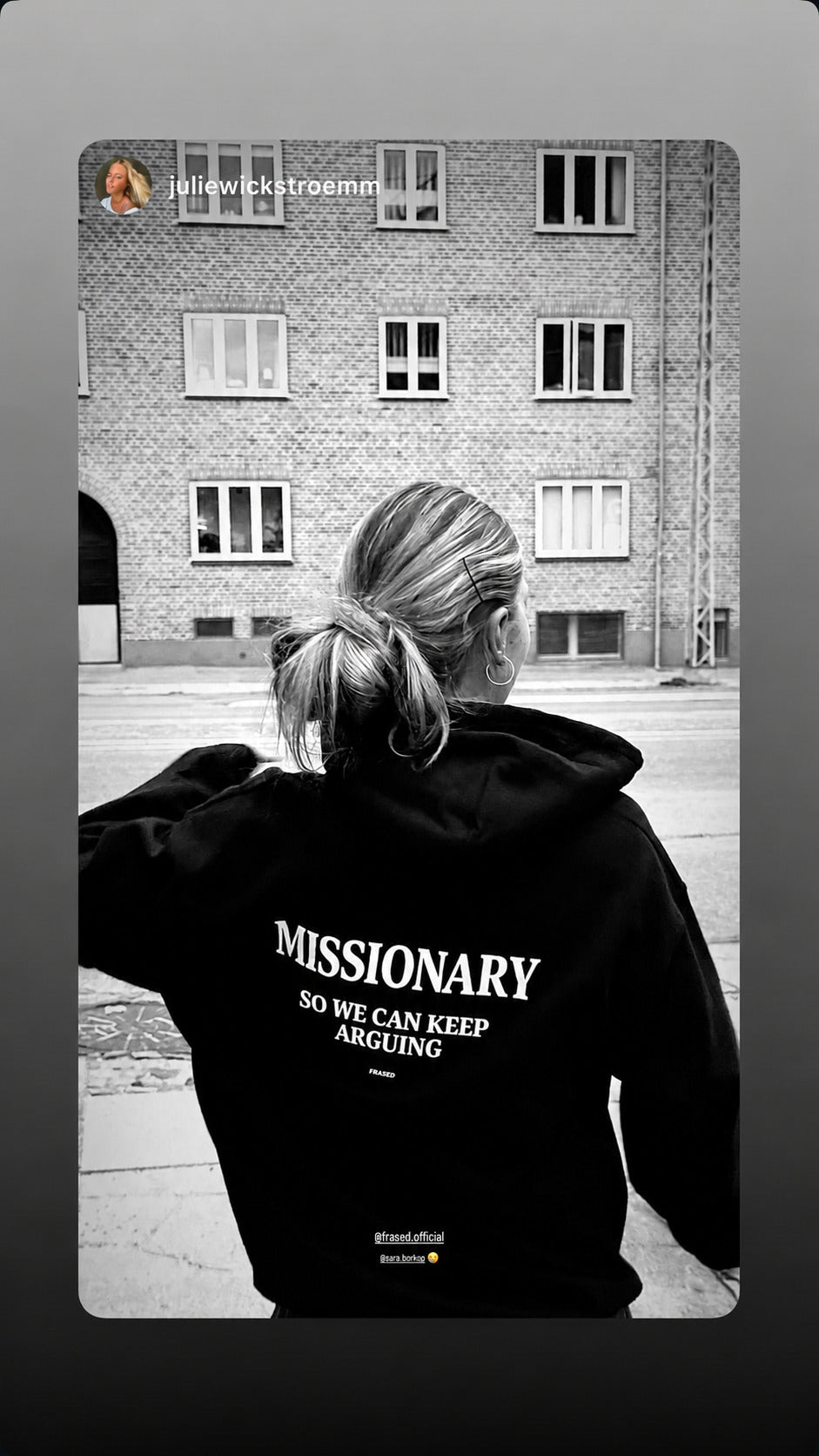 Black-and-white street photo of a woman in a black FRASED hoodie with “Missionary So We Can Keep Arguing” on the back.