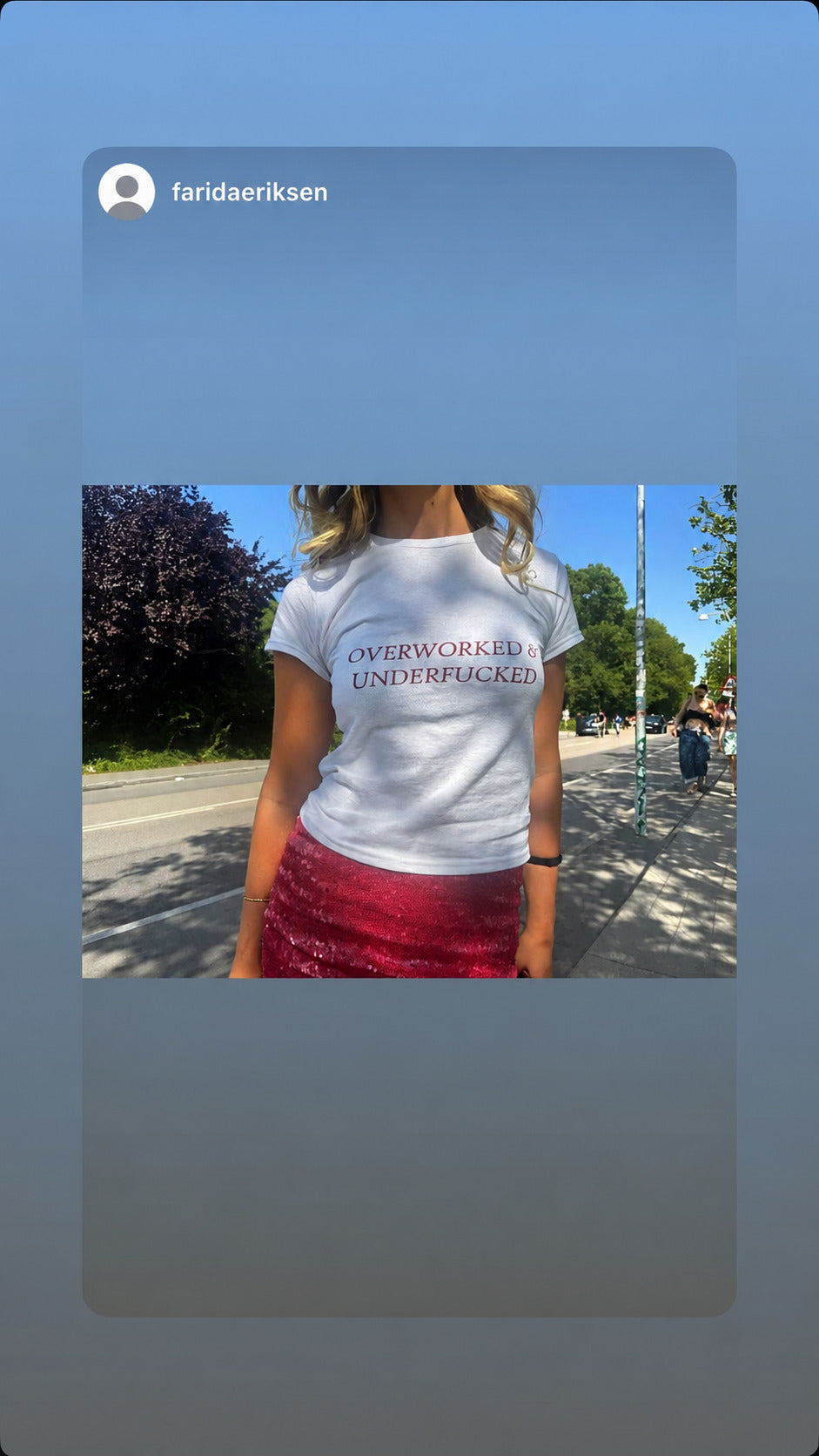 Kvinde i hvid OVERWORKED & UNDERFUCKED T-shirt og pink nederdel på en solrig gade.