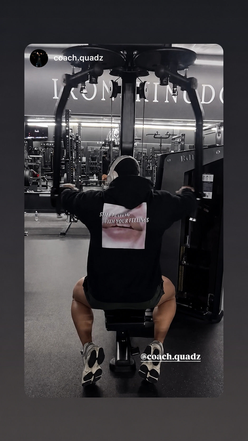 Black-and-white photo of a person in a “STILL PLAYING WITH YOUR FEELINGS” hoodie beside gym equipment.