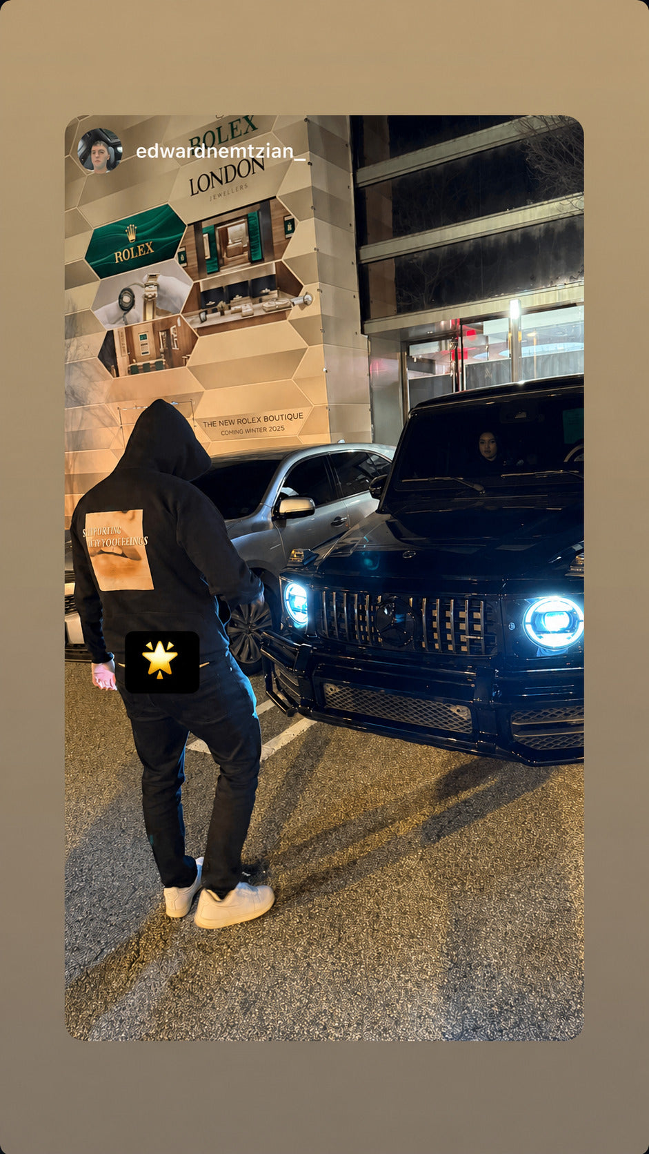 Person beside a car on a street at night in a black “STILL PLAYING WITH YOUR FEELINGS” hoodie.