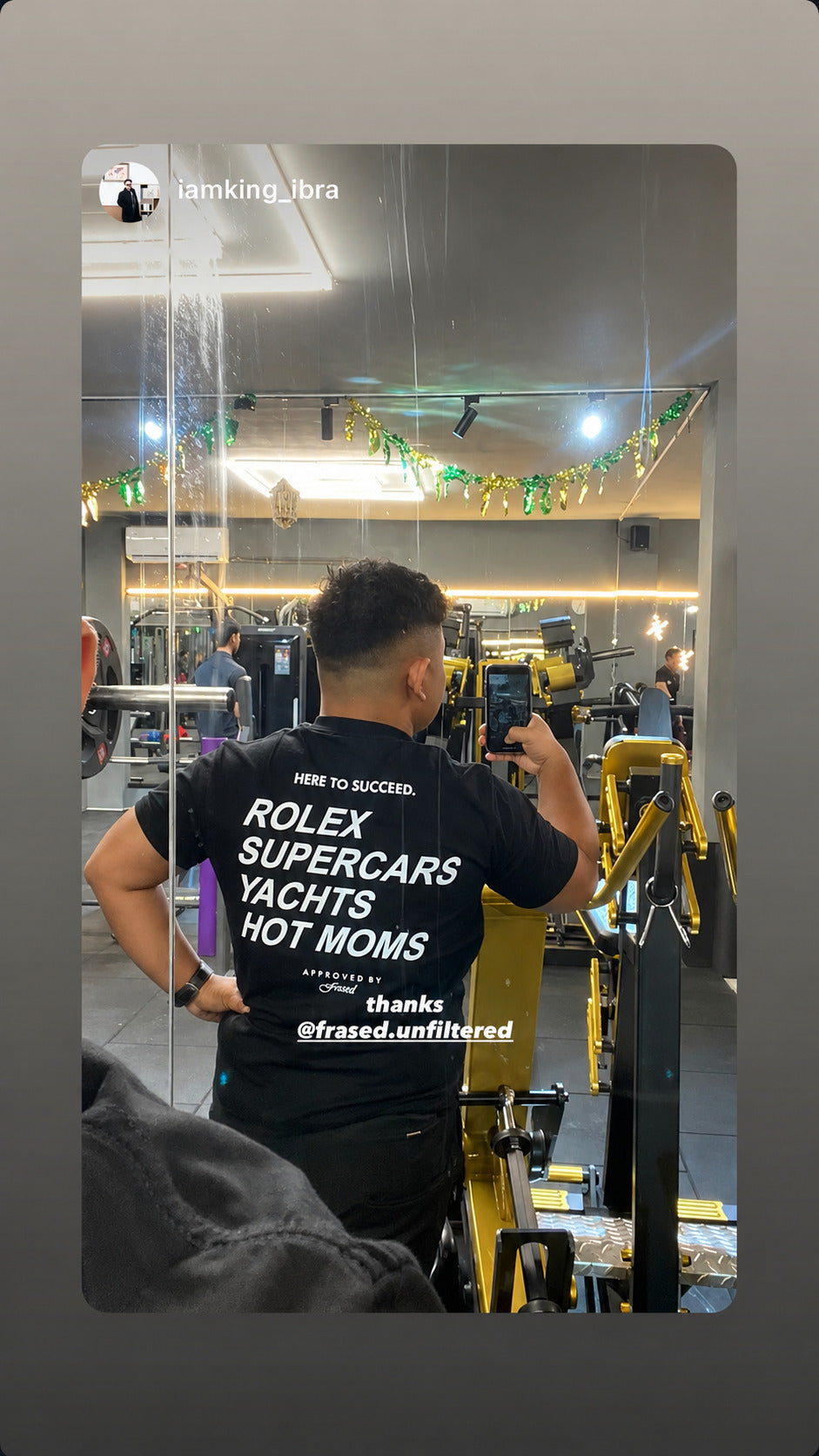 Spejlbillede i fitnesscenter af person i sort FRASED T-shirt med 'Here to succeed. Rolex Supercars Yachts Hot Moms' på ryggen.