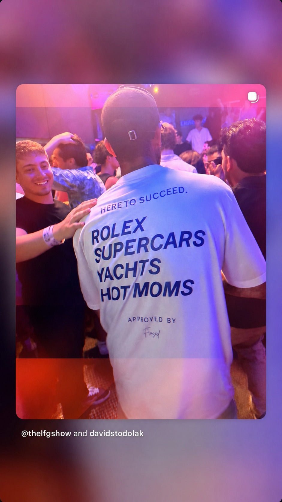 Person i klub iført hvid FRASED T-shirt med 'Here to succeed. Rolex Supercars Yachts Hot Moms' på ryggen.
