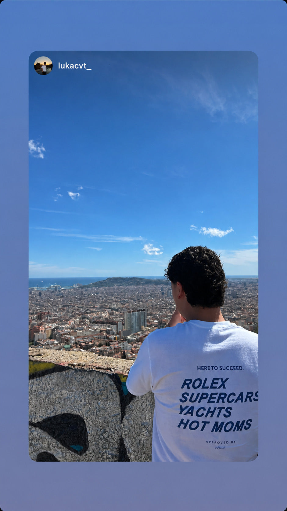 Person ved udsigt over en by iført hvid FRASED T-shirt med blåt 'Here to succeed. Rolex Supercars Yachts Hot Moms'-rygtryk.