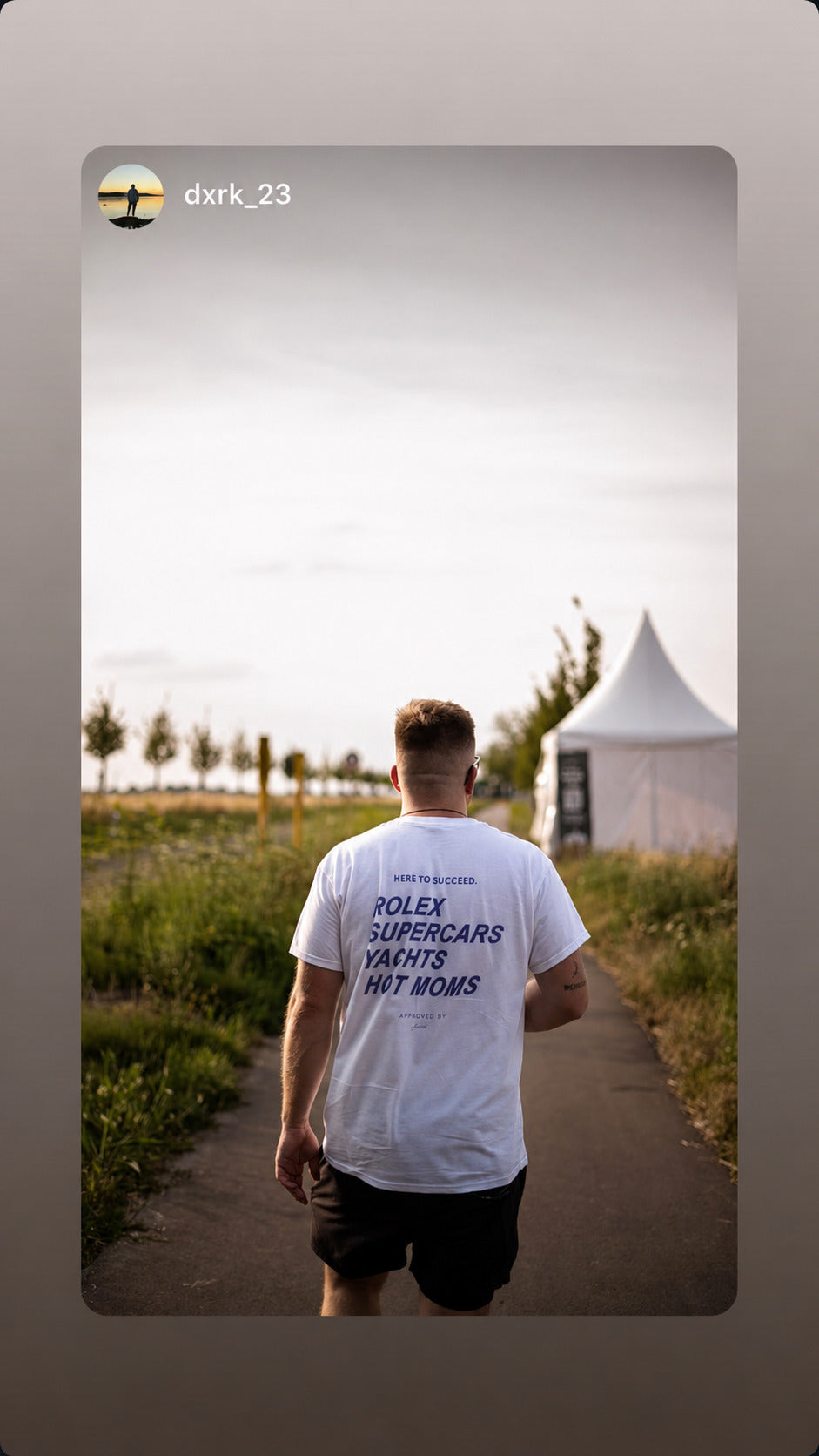 Mand går ad sti iført hvid FRASED T-shirt med 'Here to succeed. Rolex Supercars Yachts Hot Moms' på ryggen.