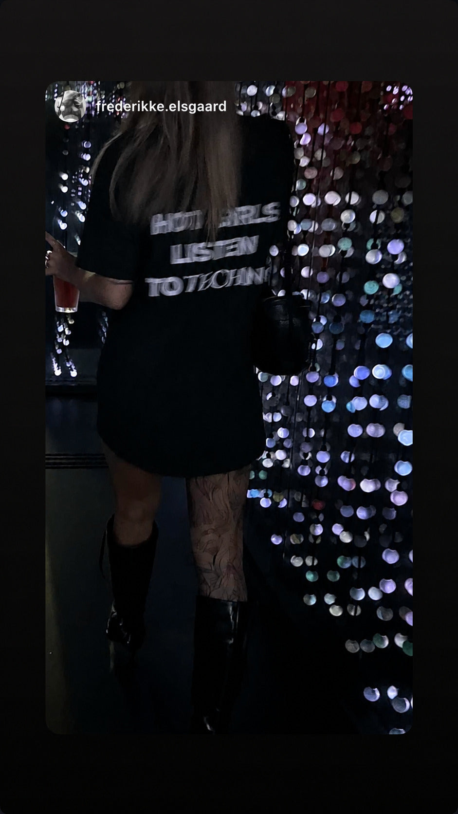 Woman seen from behind in front of a glittering wall wearing a black “HOT GIRLS LISTEN TO TECHNO” T-shirt.