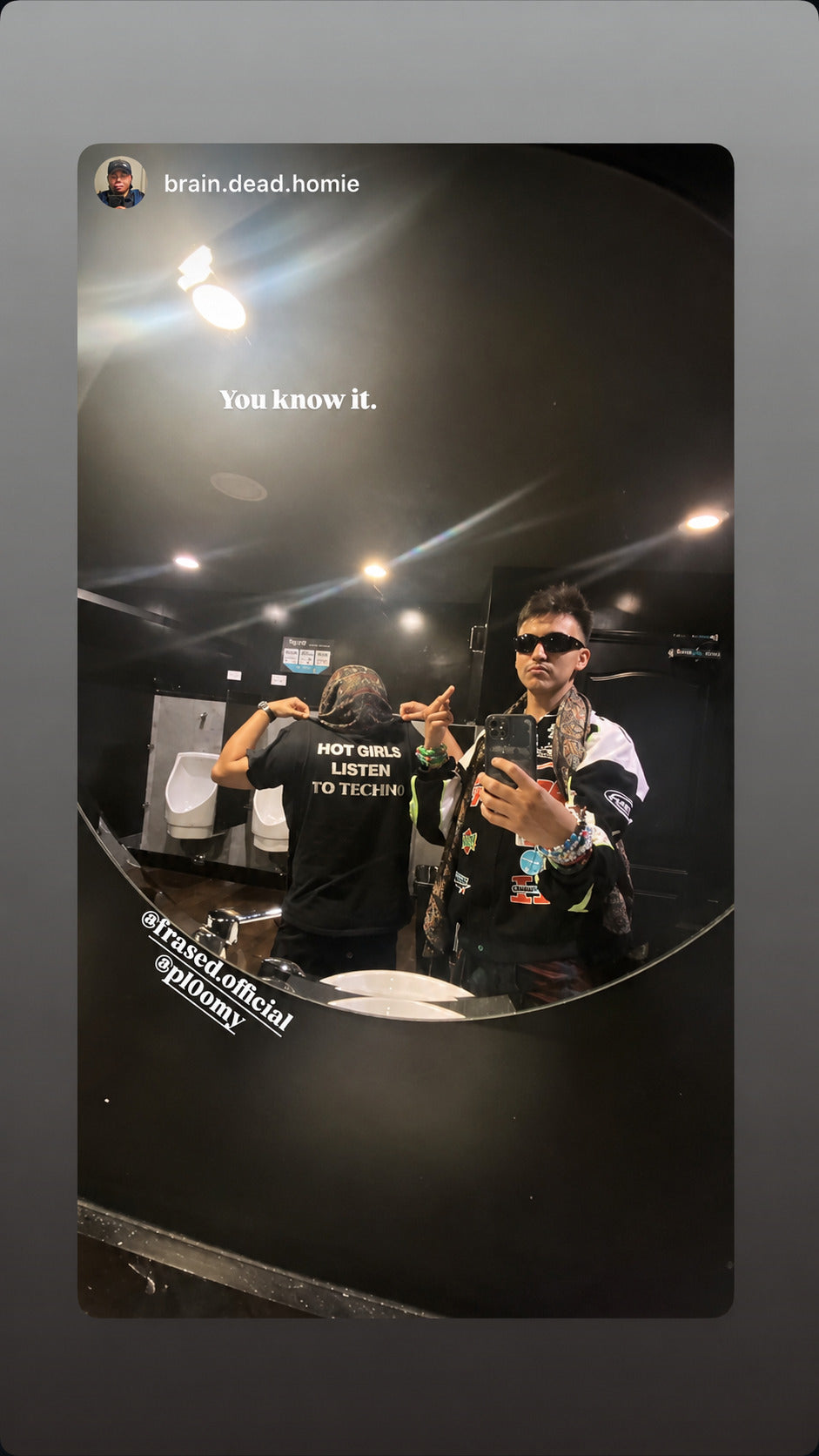 Two people taking a mirror selfie; one shows the back of a black “HOT GIRLS LISTEN TO TECHNO” T-shirt.