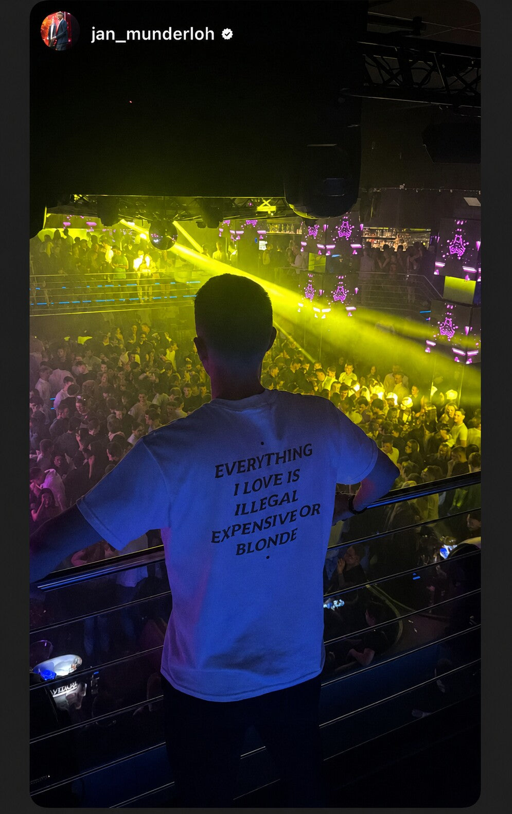 Man in a white “EVERYTHING I LOVE IS ILLEGAL EXPENSIVE OR BLONDE” T-shirt looking out over a crowded dance floor from a balcony.