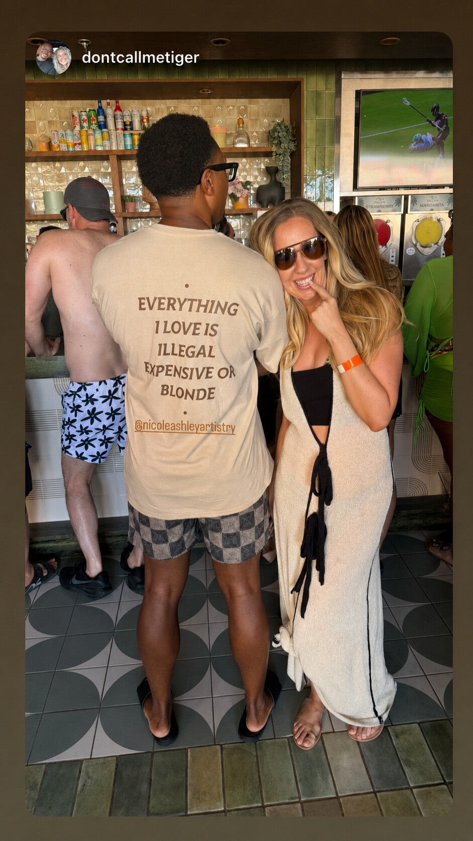 Man at a bar with a woman wearing a beige “EVERYTHING I LOVE IS ILLEGAL EXPENSIVE OR BLONDE” T-shirt.
