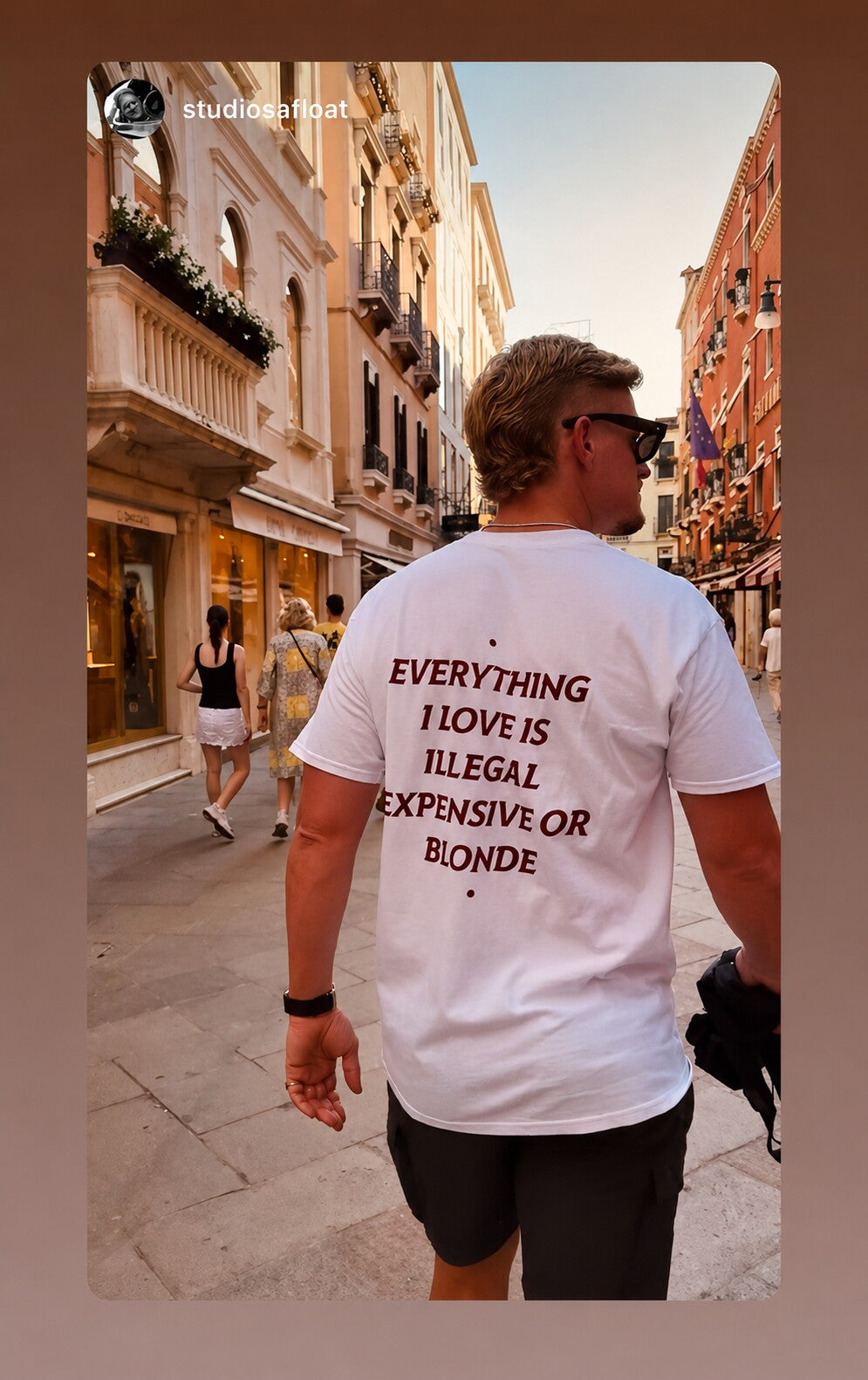 Man walking down a sunlit city street in a white “EVERYTHING I LOVE IS ILLEGAL EXPENSIVE OR BLONDE” T-shirt.