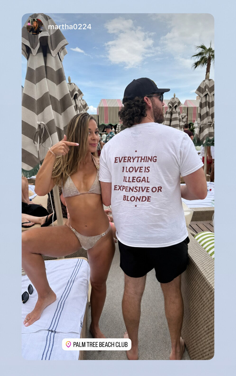 Man in a white “EVERYTHING I LOVE IS ILLEGAL EXPENSIVE OR BLONDE” T-shirt at a beach club; a woman points to the chest print.