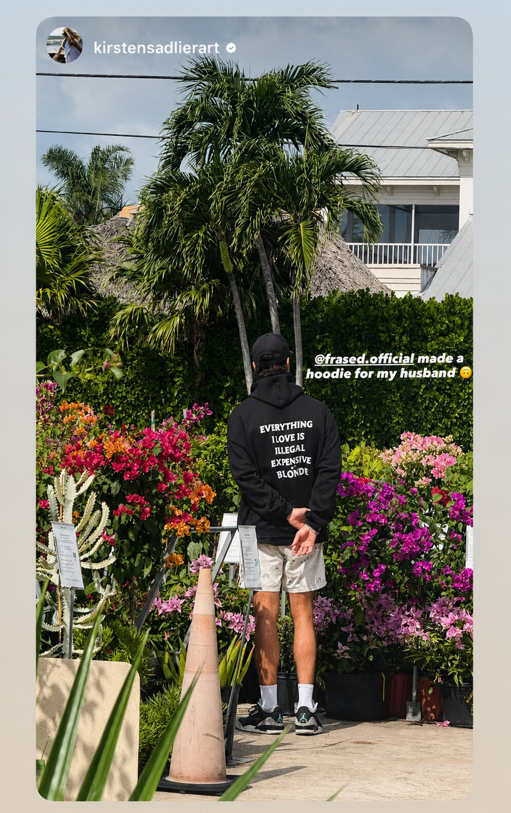 Man among flowering shrubs and palms in a black hoodie with “EVERYTHING I LOVE IS ILLEGAL EXPENSIVE OR BLONDE” on the back.