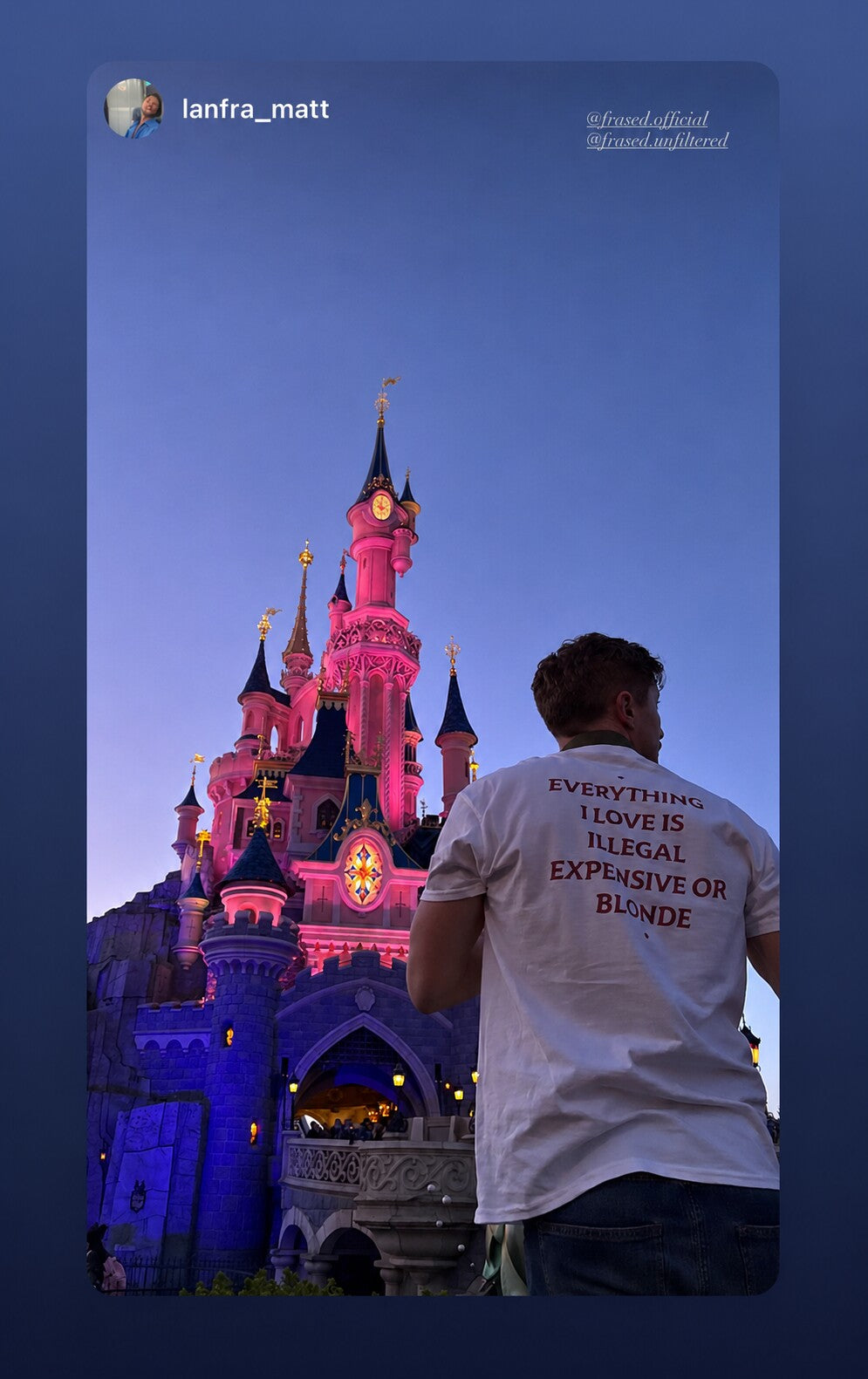 Man in a white “EVERYTHING I LOVE IS ILLEGAL EXPENSIVE OR BLONDE” T-shirt in front of an illuminated castle.