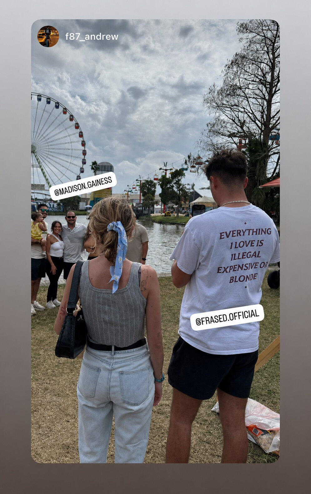 Man by a Ferris wheel in a white “EVERYTHING I LOVE IS ILLEGAL EXPENSIVE OR BLONDE” T-shirt.