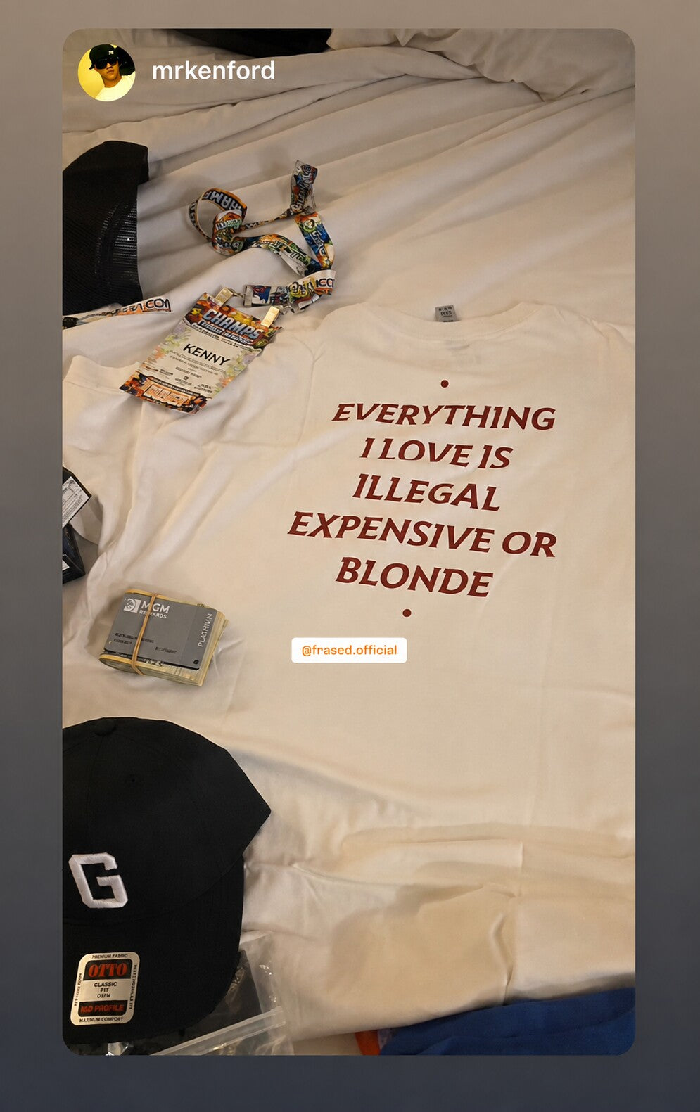 White “EVERYTHING I LOVE IS ILLEGAL EXPENSIVE OR BLONDE” T-shirt laid on a bed beside an accreditation pass.