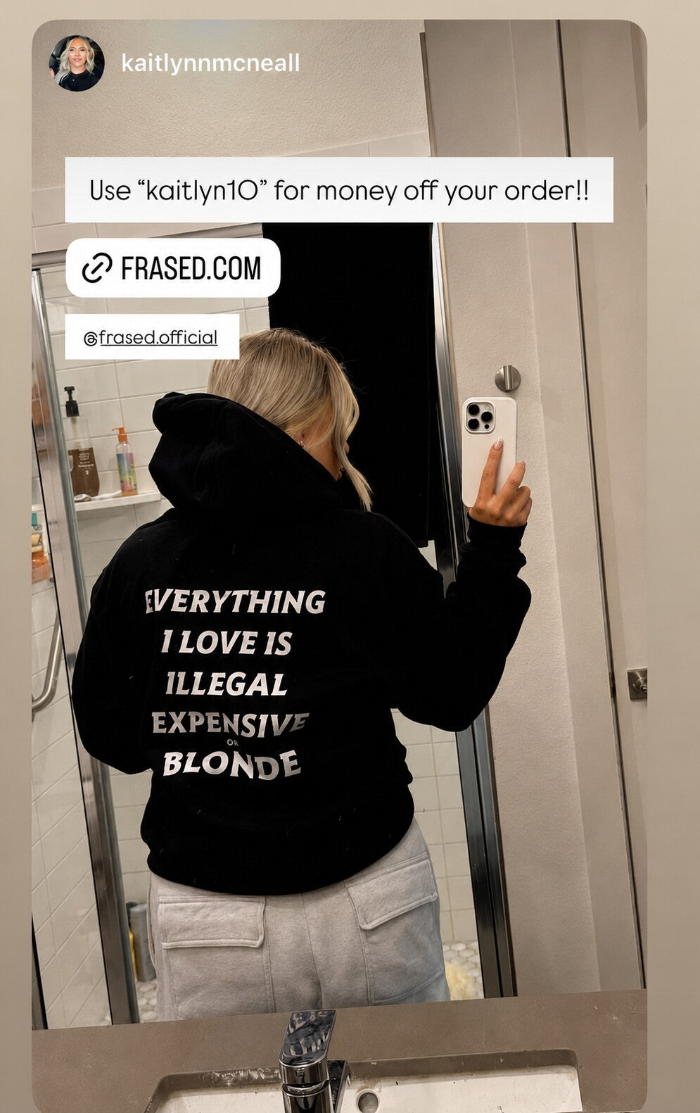 Woman taking a bathroom mirror selfie in a black hoodie with white “EVERYTHING I LOVE IS ILLEGAL EXPENSIVE OR BLONDE” back print.