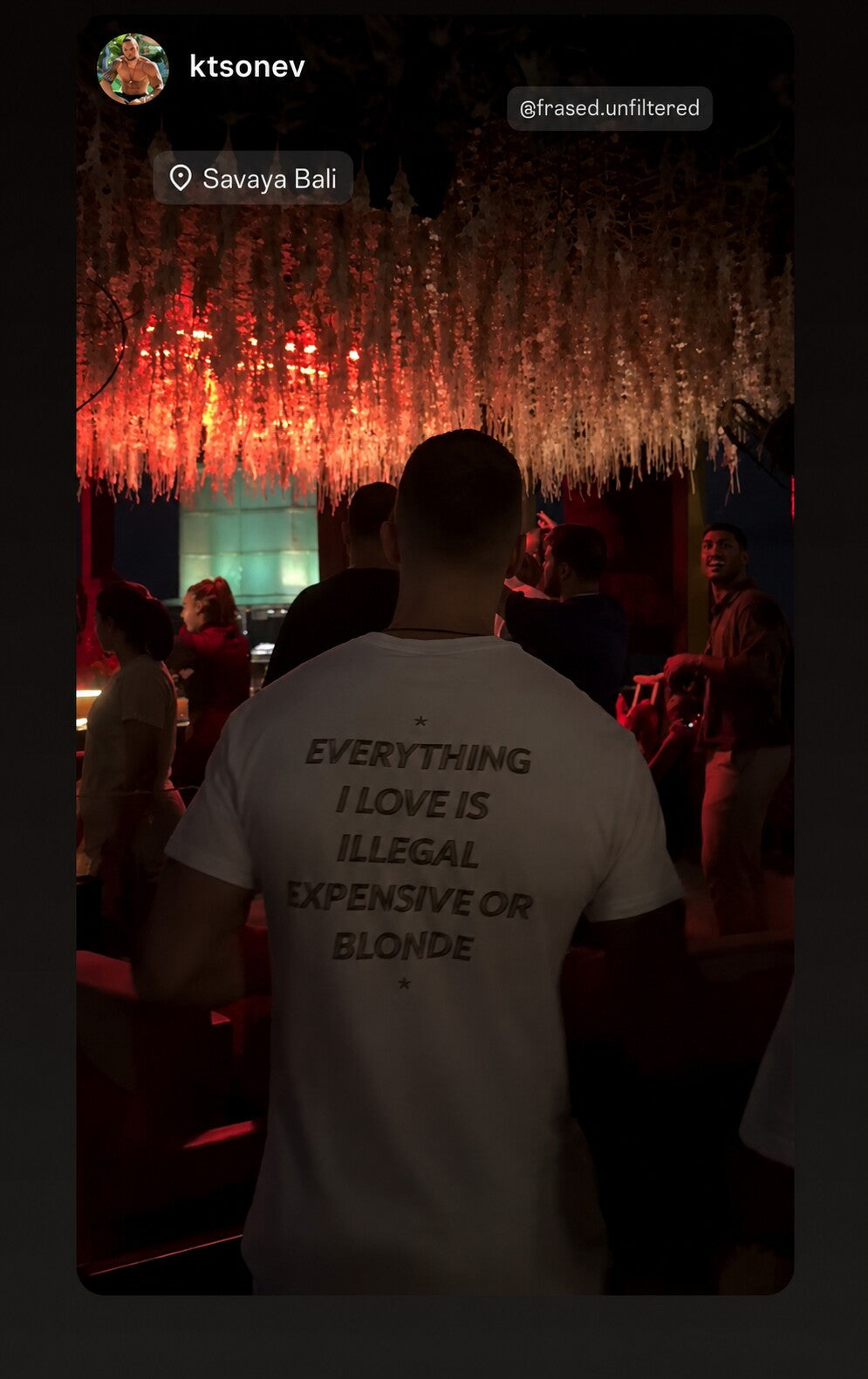 Person in a nightclub wearing a white “EVERYTHING I LOVE IS ILLEGAL EXPENSIVE OR BLONDE” T-shirt.