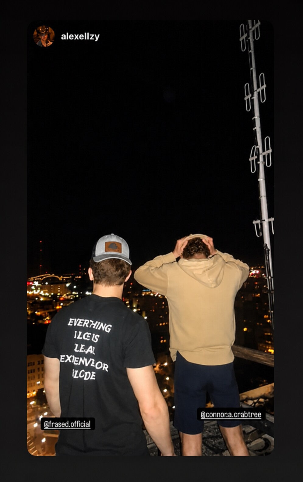 Two people overlooking city lights; one wears a black “EVERYTHING I LOVE IS ILLEGAL EXPENSIVE OR BLONDE” T-shirt.