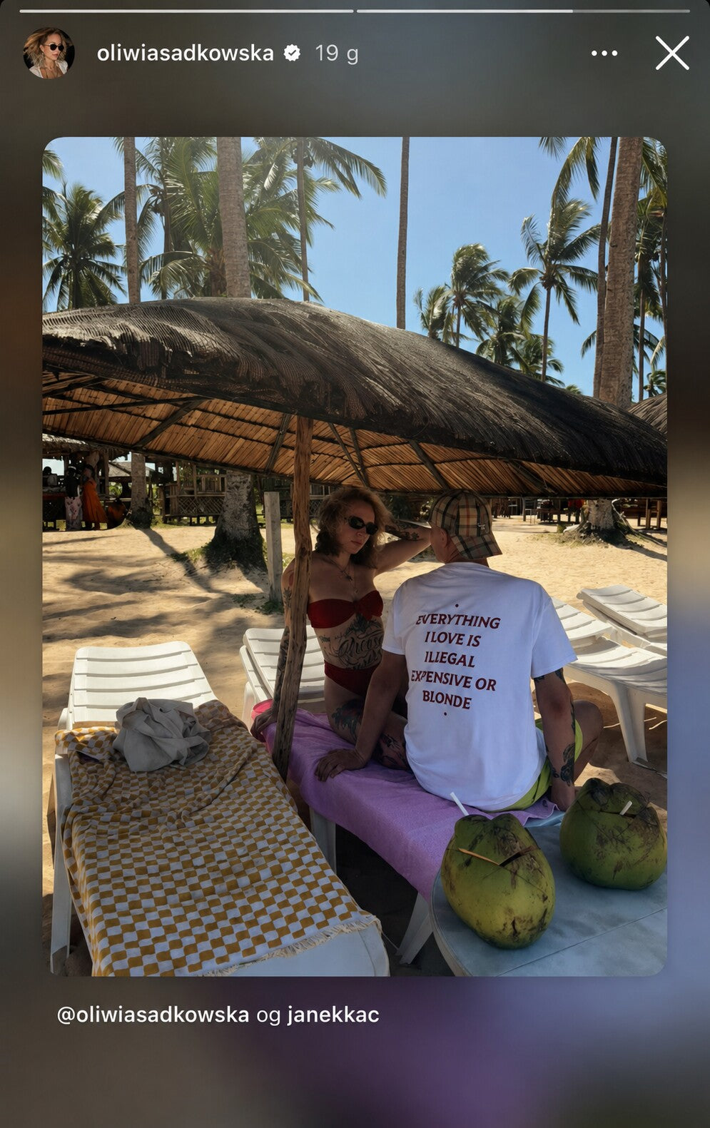 Couple beside beach chairs; the man wears a white “EVERYTHING I LOVE IS ILLEGAL EXPENSIVE OR BLONDE” T-shirt.
