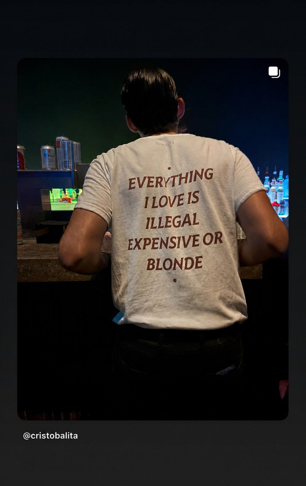 Man at a bar counter wearing a white “EVERYTHING I LOVE IS ILLEGAL EXPENSIVE OR BLONDE” T-shirt.
