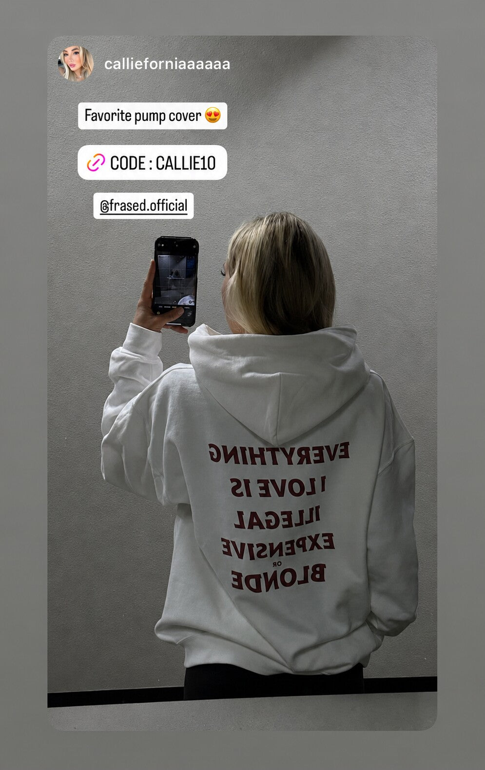 Person taking a mirror selfie in a white hoodie with “EVERYTHING I LOVE IS ILLEGAL EXPENSIVE OR BLONDE” on the back, reversed in the reflection.