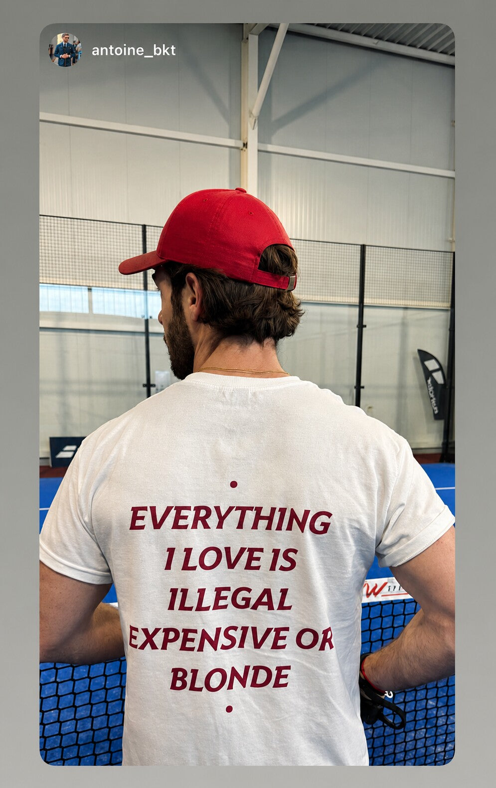 Man beside an indoor court wearing a white “EVERYTHING I LOVE IS ILLEGAL EXPENSIVE OR BLONDE” T-shirt.