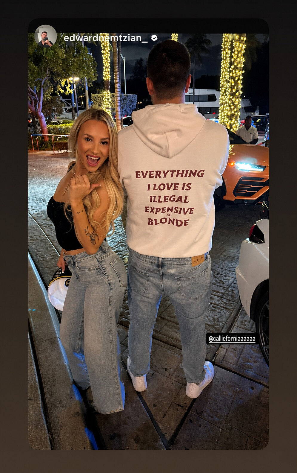 Man and woman on a lit street at night; he shows the “EVERYTHING I LOVE IS ILLEGAL EXPENSIVE OR BLONDE” back print on a white hoodie.
