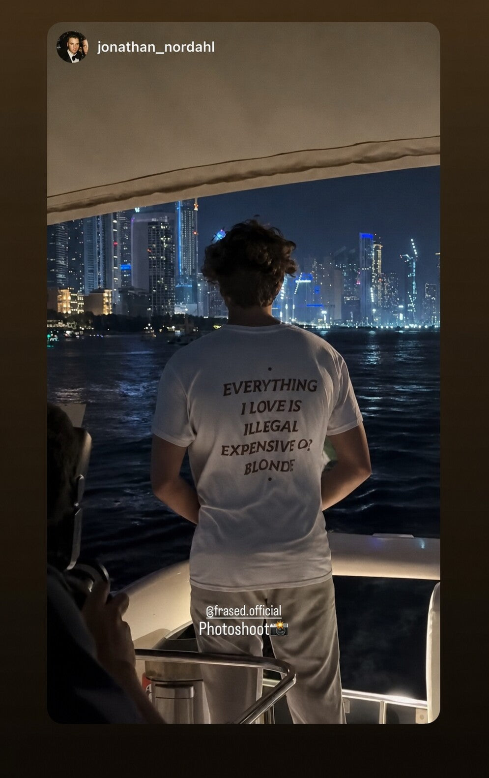 Man on a boat with city lights behind him wearing a white “EVERYTHING I LOVE IS ILLEGAL EXPENSIVE OR BLONDE” T-shirt.