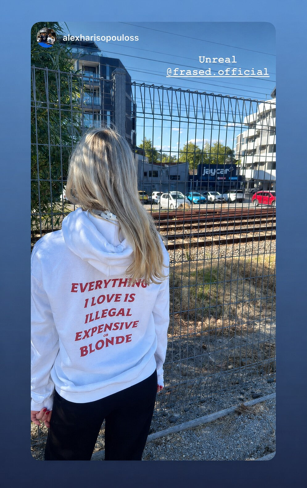 Blonde person beside a railway fence, seen from behind in a white hoodie with red “EVERYTHING I LOVE IS ILLEGAL EXPENSIVE OR BLONDE” back print.
