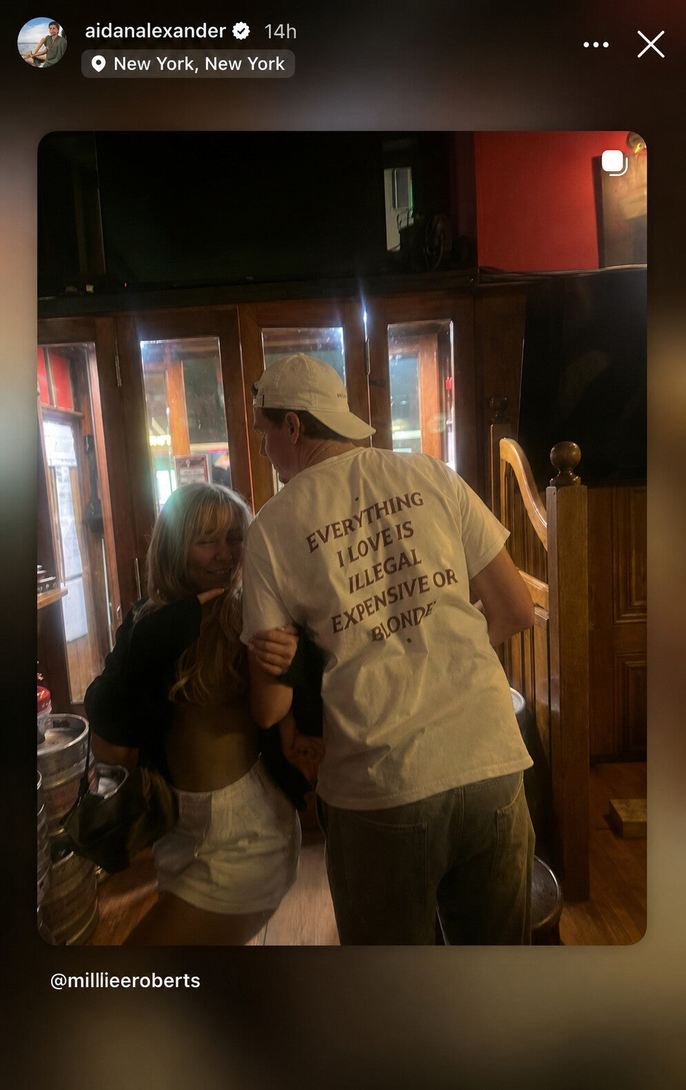 Man with a woman at a bar wearing a white “EVERYTHING I LOVE IS ILLEGAL EXPENSIVE OR BLONDE” T-shirt.