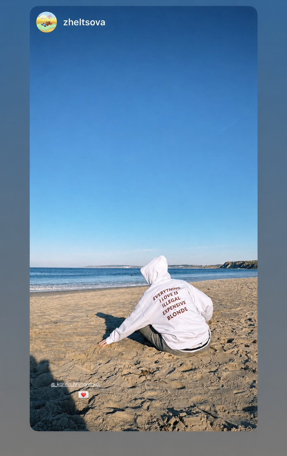 Person sitting on a sandy beach facing the sea in a white hoodie with “EVERYTHING I LOVE IS ILLEGAL EXPENSIVE OR BLONDE” on the back.