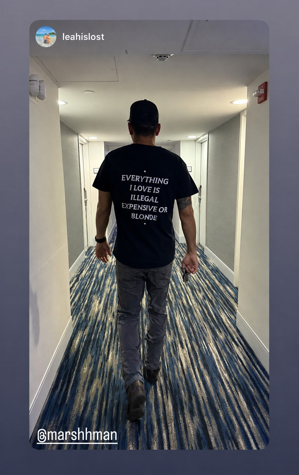 Man in a hallway wearing a black “EVERYTHING I LOVE IS ILLEGAL EXPENSIVE OR BLONDE” T-shirt.