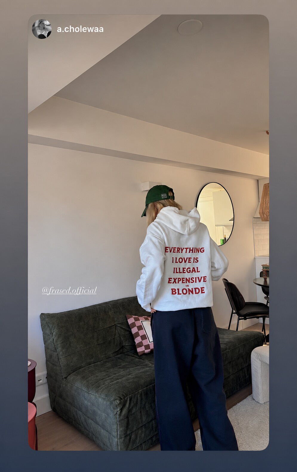 Person in a living room wearing a green cap, seen from behind in a white hoodie with red “EVERYTHING I LOVE IS ILLEGAL EXPENSIVE OR BLONDE” print.