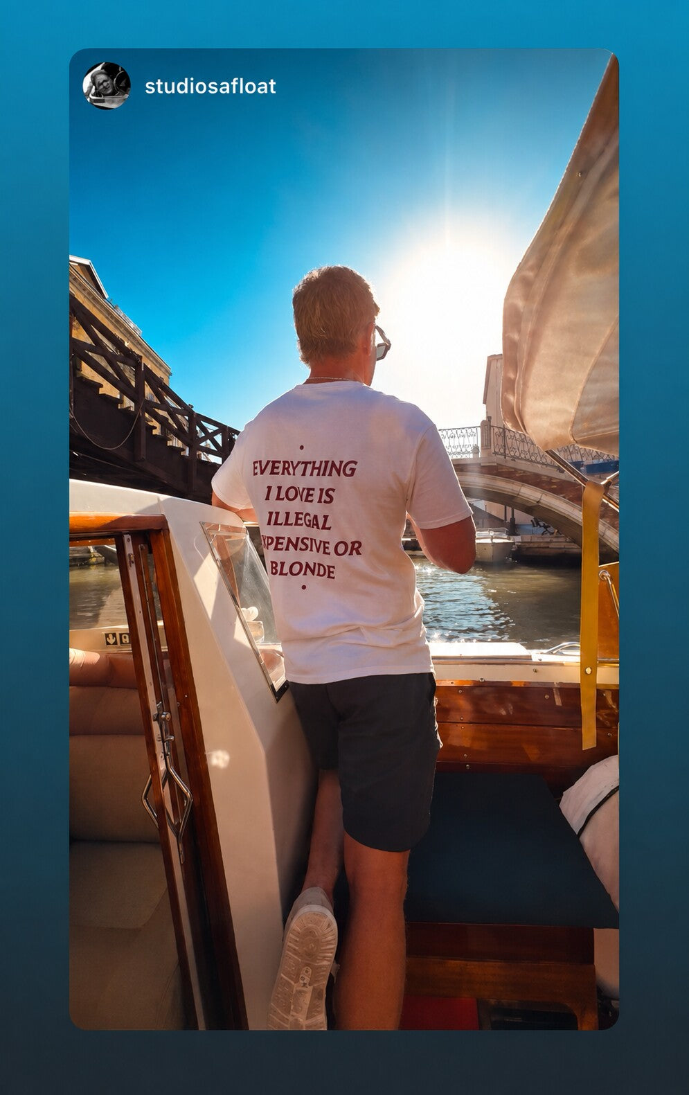 Man on a boat in sunlight wearing a white “EVERYTHING I LOVE IS ILLEGAL EXPENSIVE OR BLONDE” T-shirt.