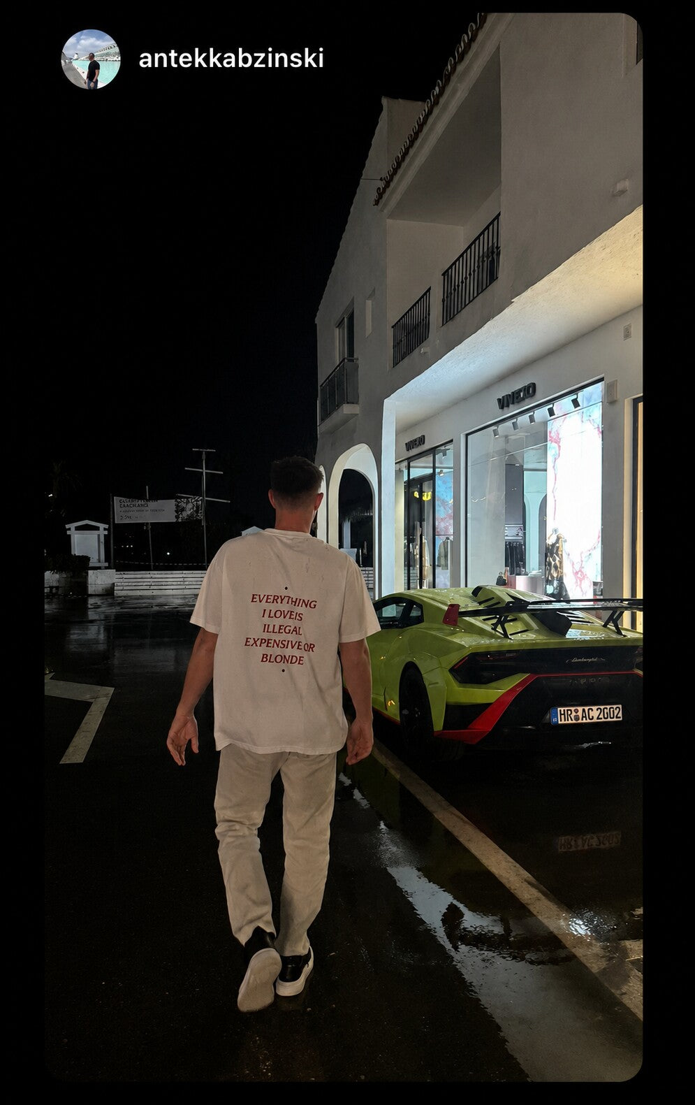 Man walking past a green sports car at night in a white “EVERYTHING I LOVE IS ILLEGAL EXPENSIVE OR BLONDE” T-shirt.